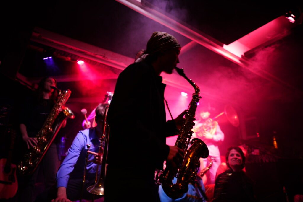Photograph of a brass band at a concert, a backlit alto saxophone player is in focus in the foreground